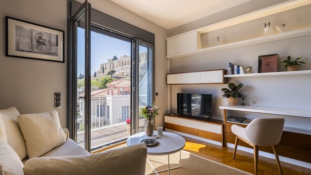 A modern living room with a sofa, a coffee table, and a TV. There is a large window with a view of a cityscape. - Phos, an eclectic suite with stunning Acropolis view.<br> (Athina)