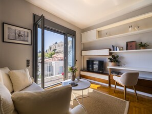 A modern living room with a sofa, a coffee table, and a TV. There is a large window with a view of a cityscape. - Phos, an eclectic suite with stunning Acropolis view.<br> (Athina)