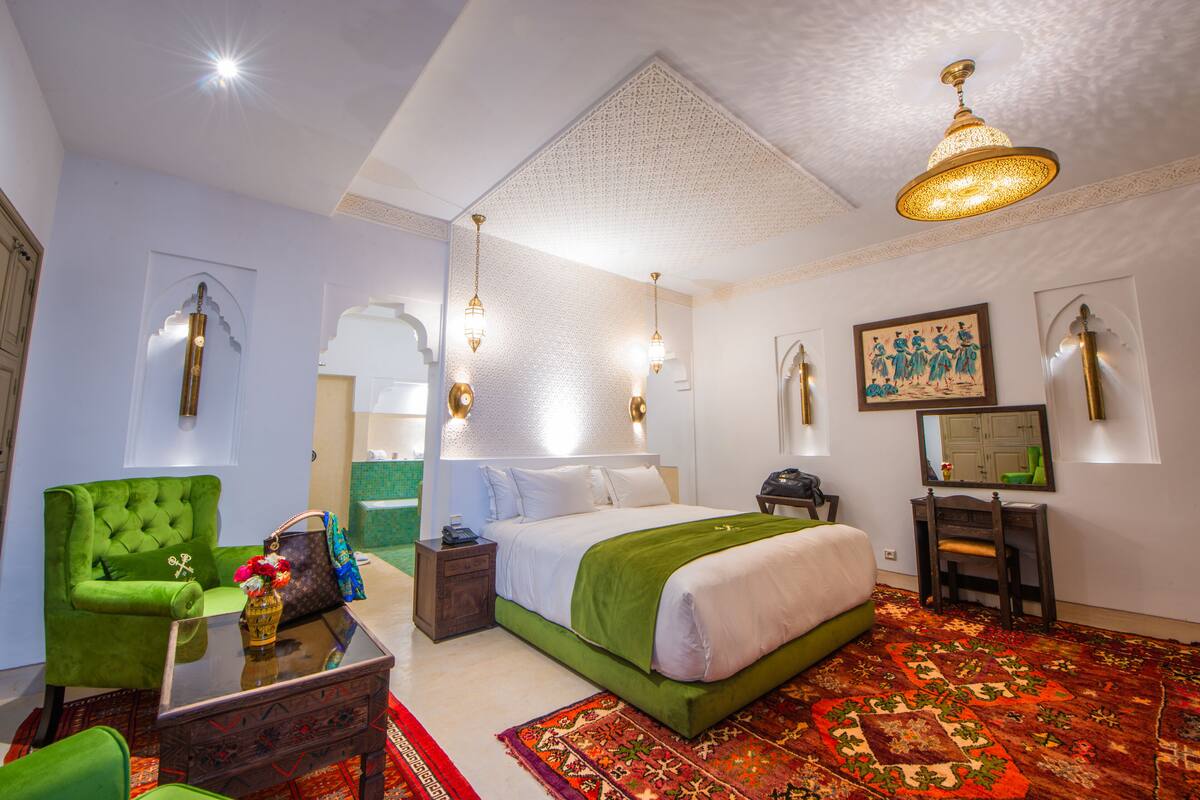 A hotel room with a green upholstered armchair, a glass coffee table, a bed with a green bedspread, a patterned rug, and a large mirror on the wall.