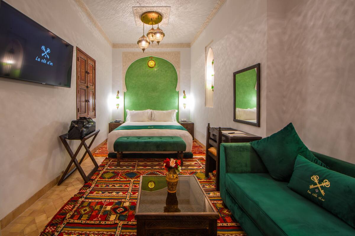 A hotel room with a bed, a green sofa, a small table with a vase, a television on the wall, and a patterned carpet.