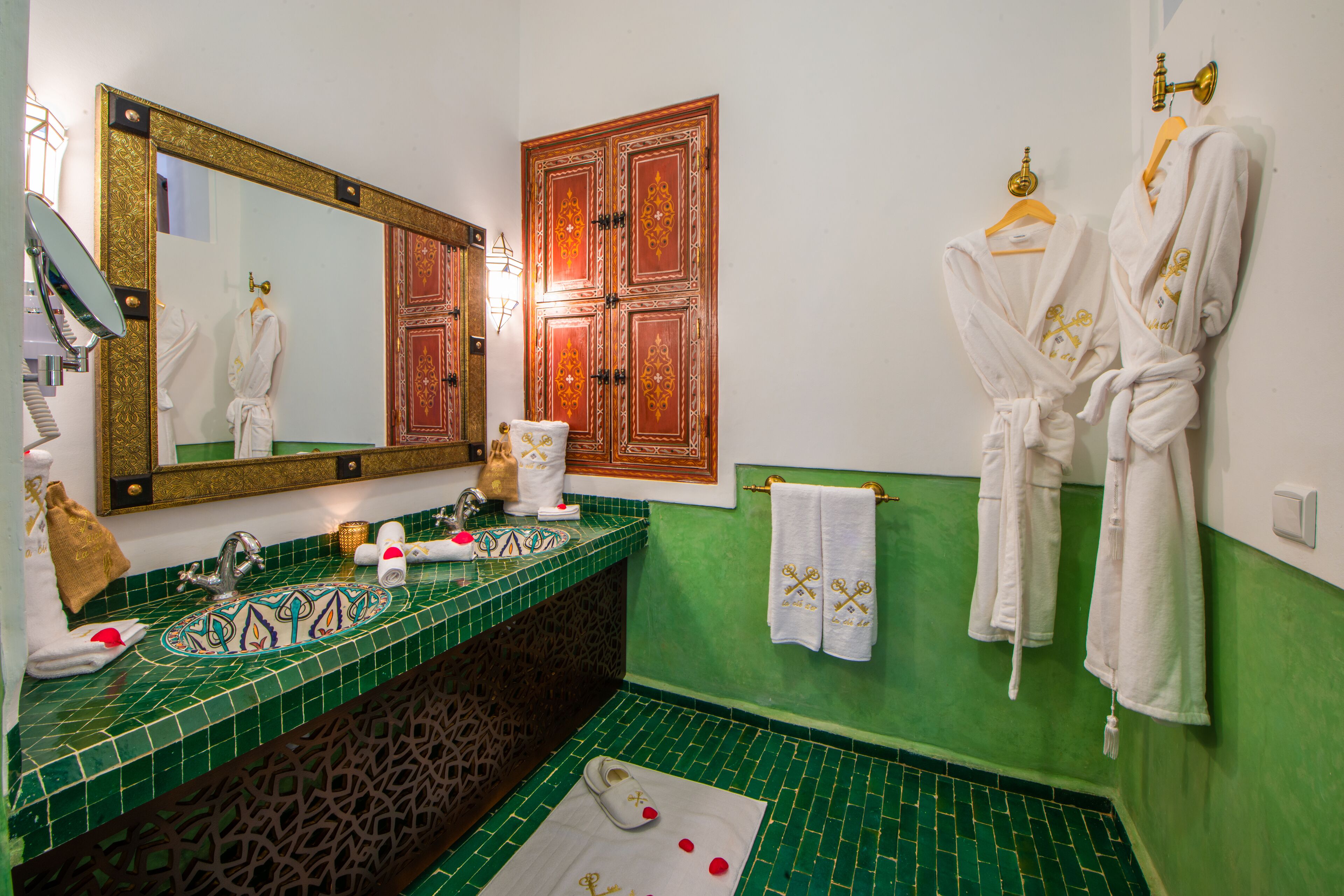 A bathroom with a green tiled sink, a large mirror, and two white robes hanging on the wall.