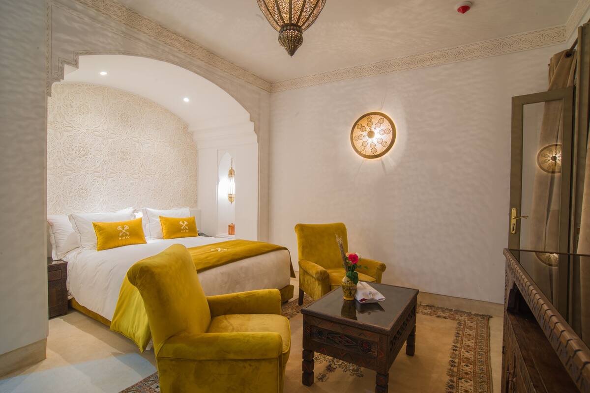 A hotel room with a bed, a yellow armchair, a small table, and a decorative wall light.