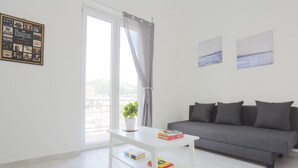 A modern living room with a sofa, a white table with books, a potted plant, and a cityscape view through a large window. - GuestHost - Bright & Central Apartment 5 min from the station (Salerno)