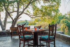A wooden dining table with chairs, set outdoors with a view of a body of water and trees.