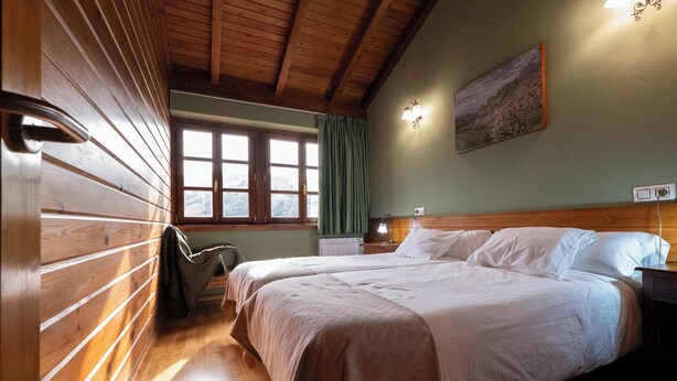 A room with wooden paneling, a large bed with white linens, a nightstand, a lamp, and a window with curtains.