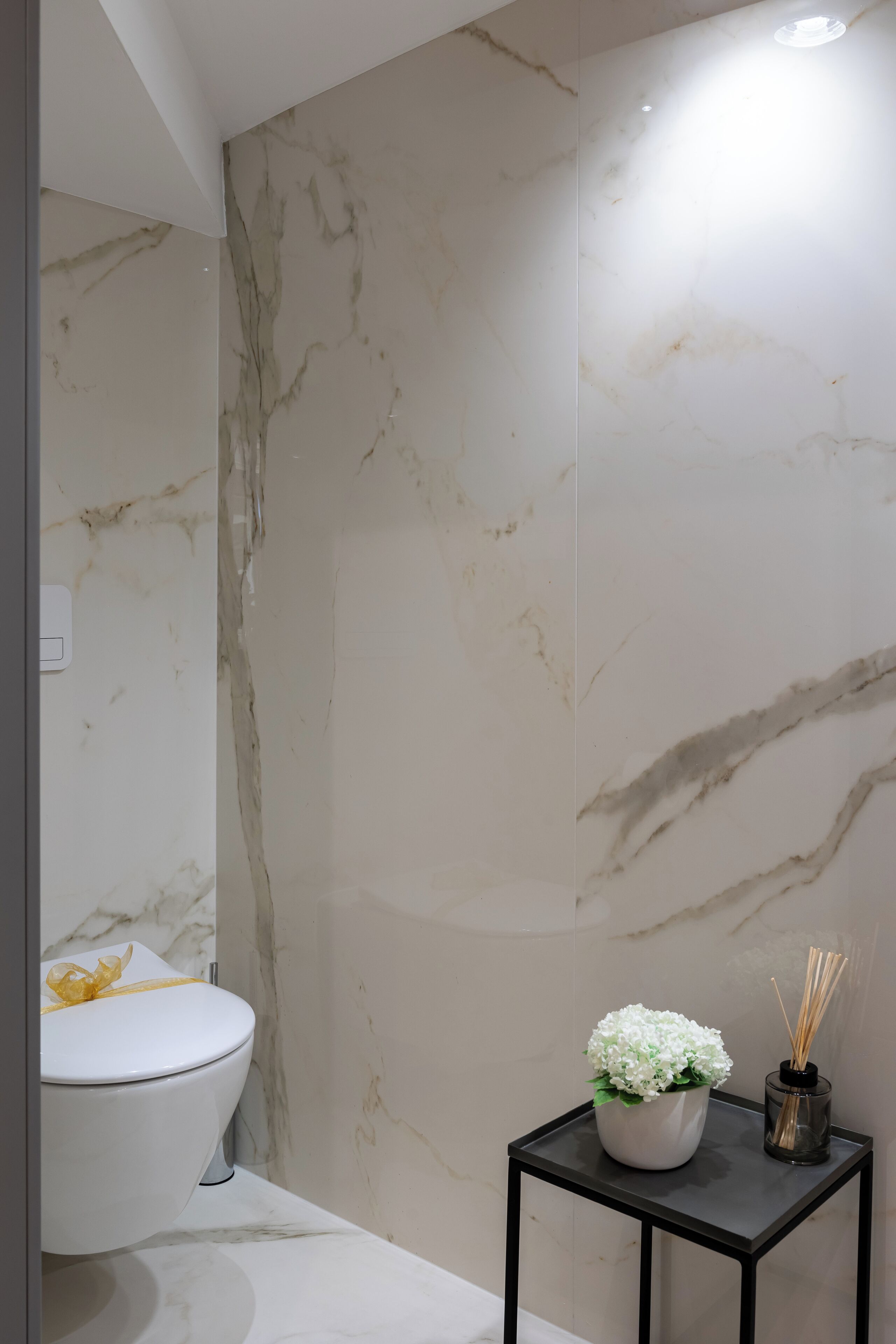 Ein Badezimmer mit einer weißen Toilette, Marmorwänden und einem kleinen schwarzen Tisch mit einer weißen Blumenvase und einem Schilferoher Diffusor.