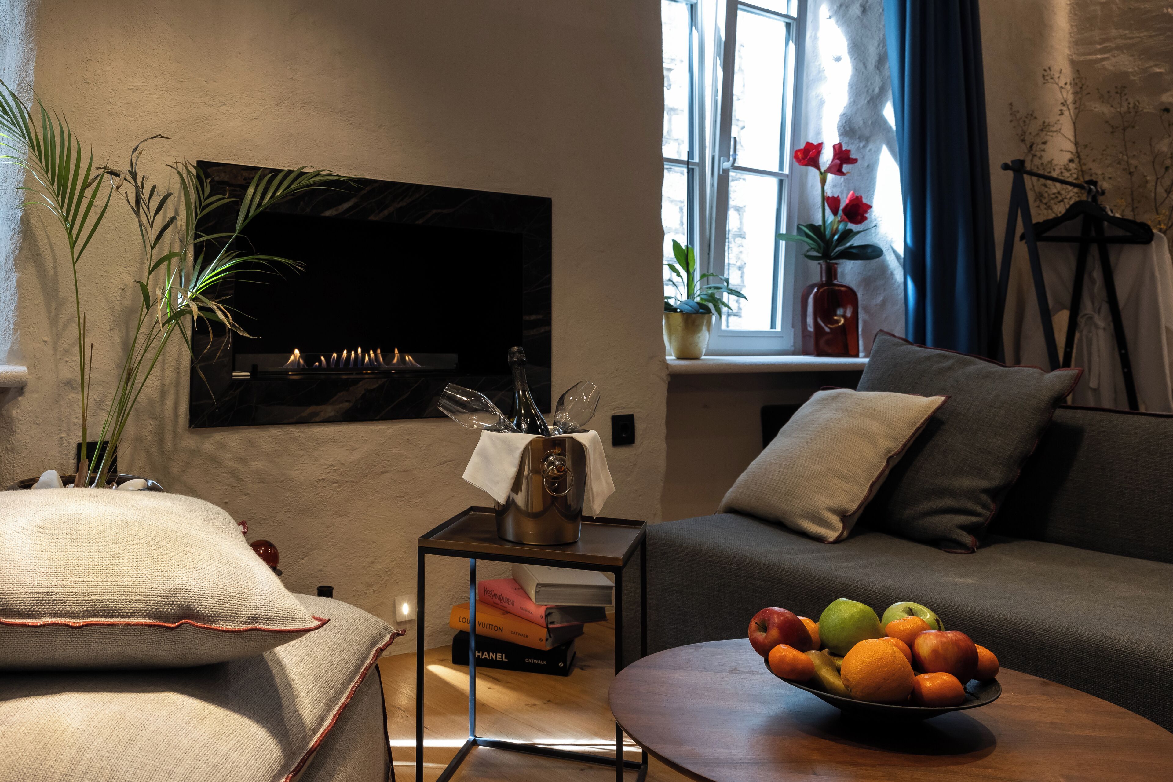 Una sala de estar con un televisor de pantalla plana, un sofá, una mesa de centro con fruta, una mesita auxiliar con libros y un jarrón, y una ventana con cortinas.