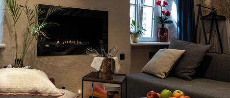 Una sala de estar con un televisor de pantalla plana, un sofá, una mesa de centro con fruta, una mesita auxiliar con libros y un jarrón, y una ventana con cortinas.