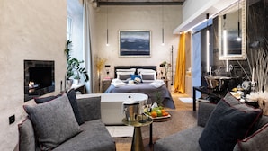 Ein modernes Hotelzimmer mit einem großen Bett, einer Sitzecke mit Sofas, einem Flachbildfernseher, einem Kamin und einem Esstisch mit Obst.