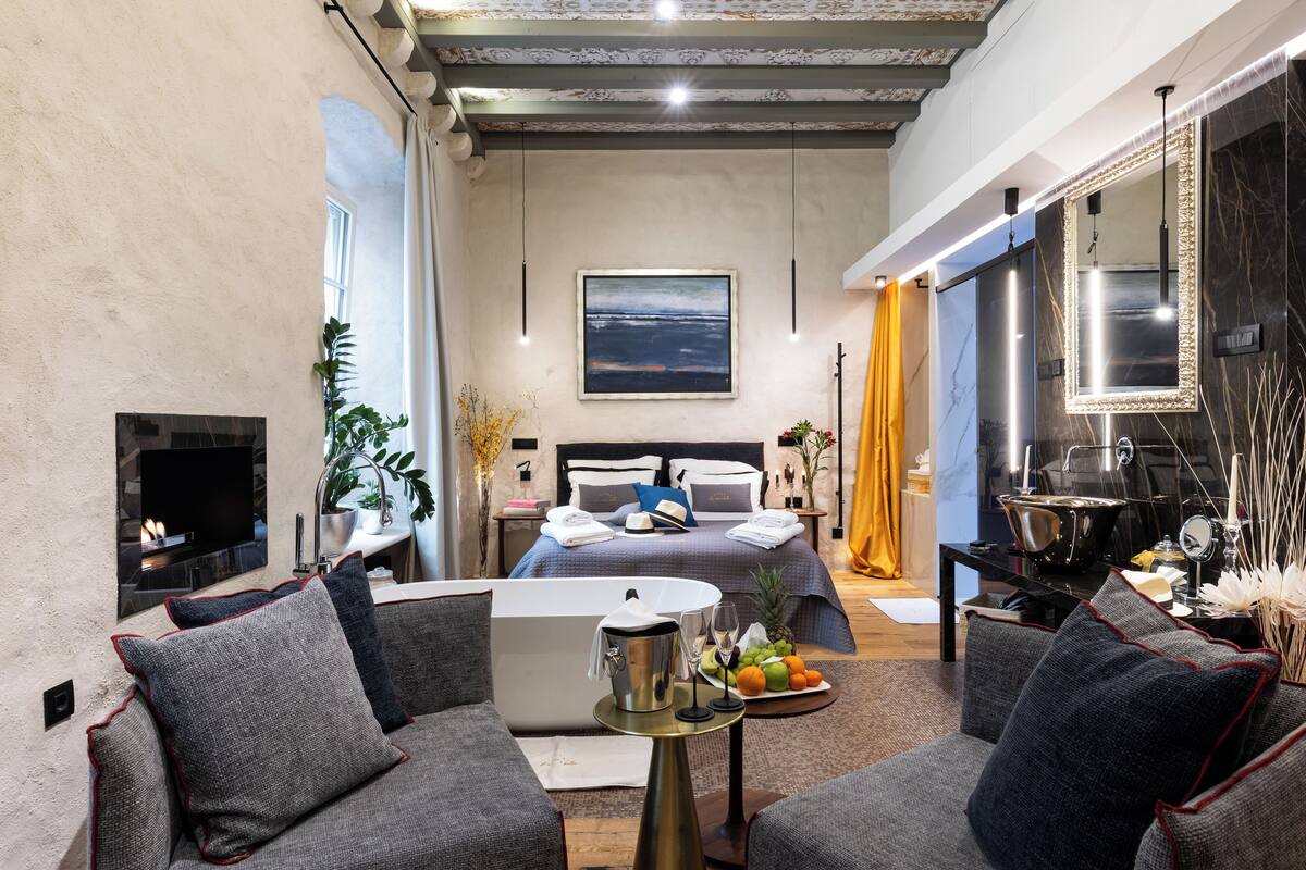 Une chambre d’hôtel moderne dotée d’un grand lit, d’un coin salon avec des canapés, d’une télévision à écran plat, d’une cheminée et d’une table à manger agrémentée de fruits.