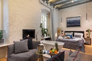 Quarto de hotel moderno com lareira, cama com vista, área de jantar com cesta de frutas e um espaço de estar com mesa de centro.