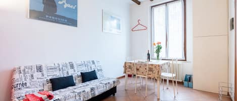 Una habitación amoblada con sofá, mesa de comedor, sillas, radiador, ventana y un cartel en la pared.