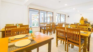 Un espace repas comprenant plusieurs tables et chaises, de grandes fenêtres offrant une vue sur les bâtiments, et un petit autel ou un objet décoratif accroché au mur.