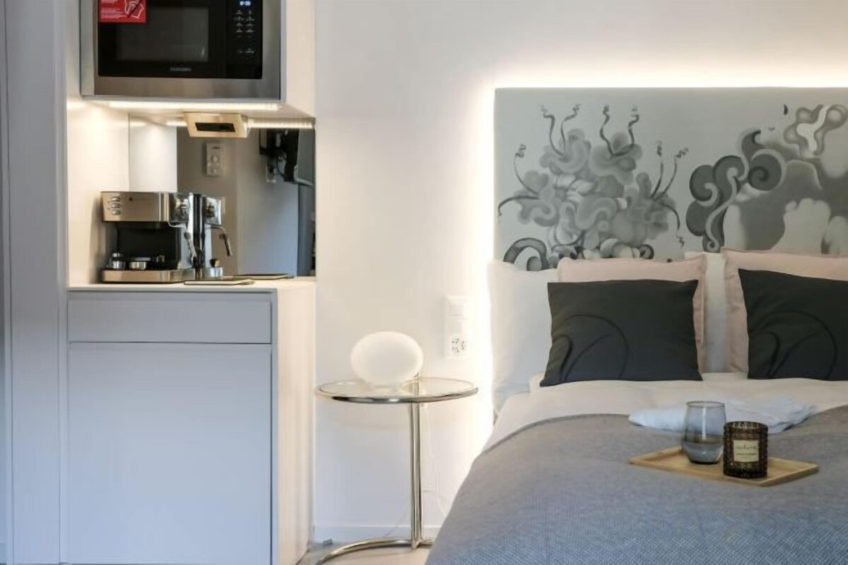 Une chambre d’hôtel compacte comprenant un coin cuisine, un lit agrémenté d’oreillers décoratifs, une table de chevet avec un verre et un plateau avec une bougie.