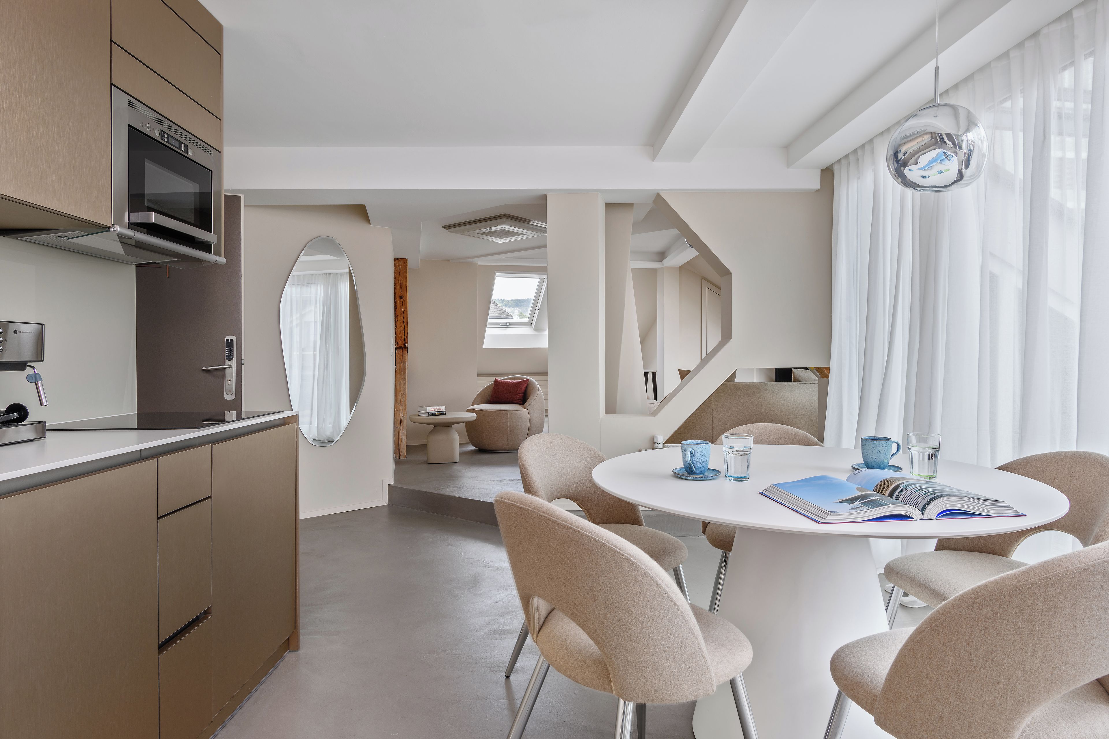 Eine moderne Küche mit eingebauten Geräten, ein Essbereich mit rundem Tisch und Stühlen sowie ein Blick durch ein Fenster.
