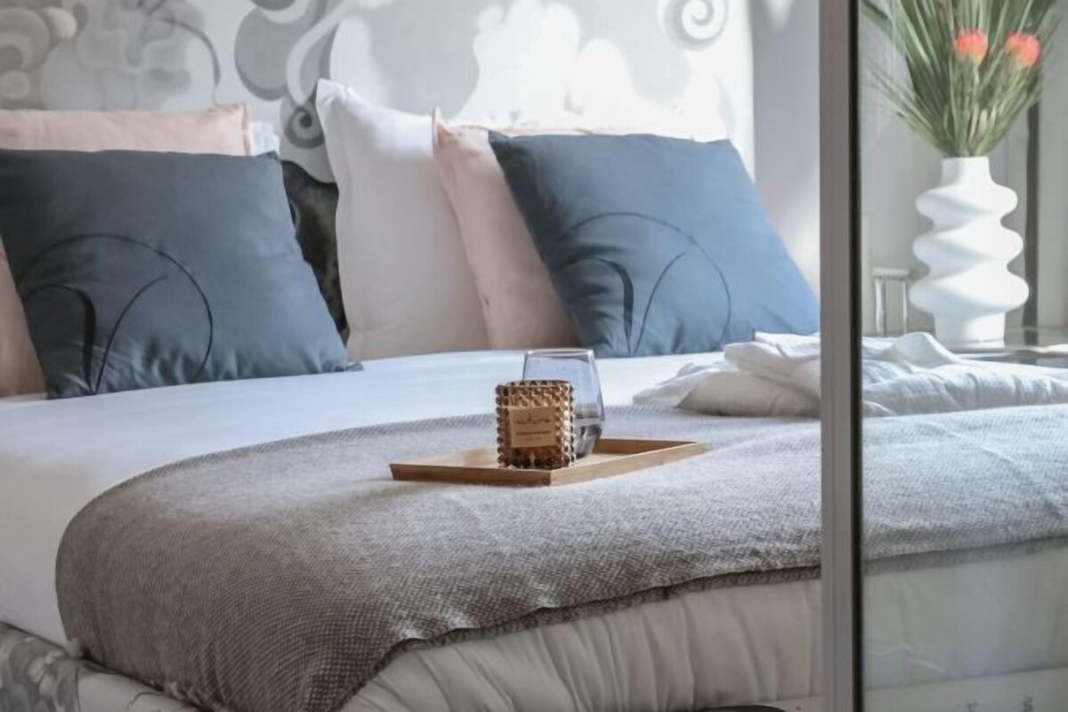 Un lit bien fait, recouvert d’une couverture grise, une table de chevet sur laquelle se trouvent un plateau avec un verre et un récipient, ainsi qu’un vase blanc orné de fleurs.