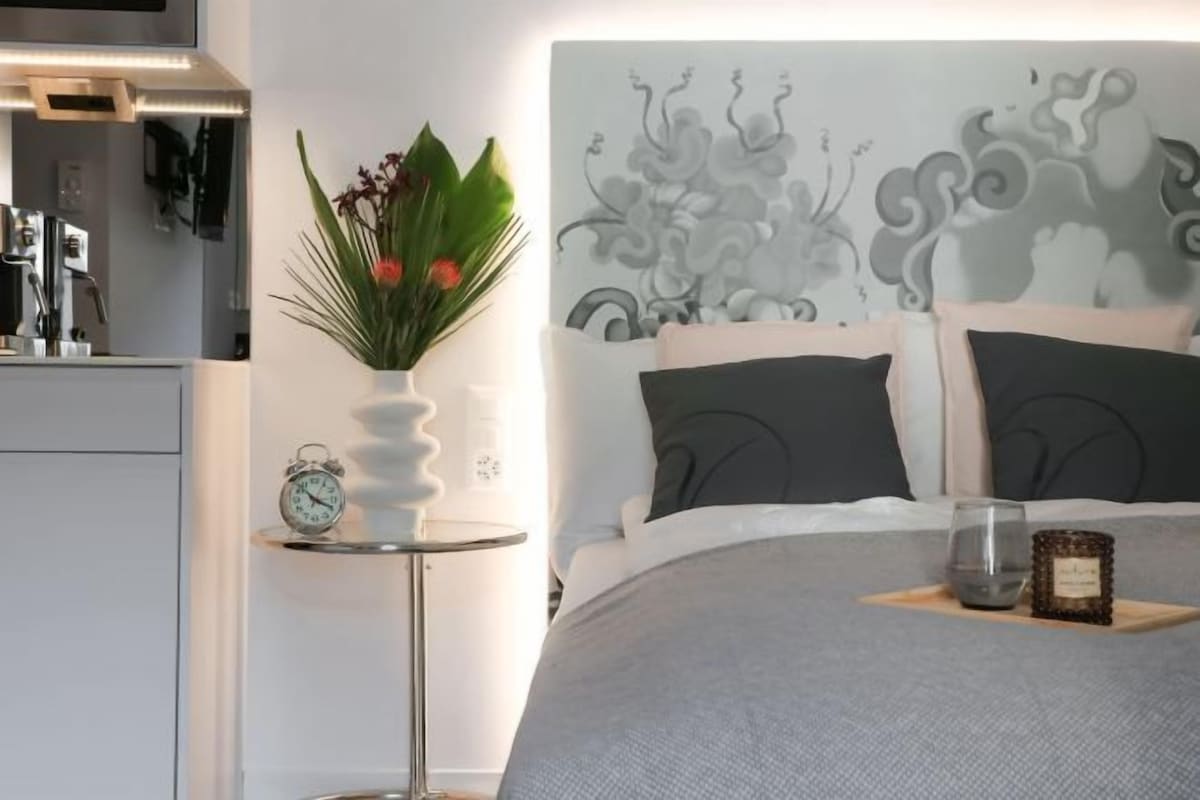 A bedroom with a bed, a bedside table with a clock, a vase with flowers, and a decorative headboard.
