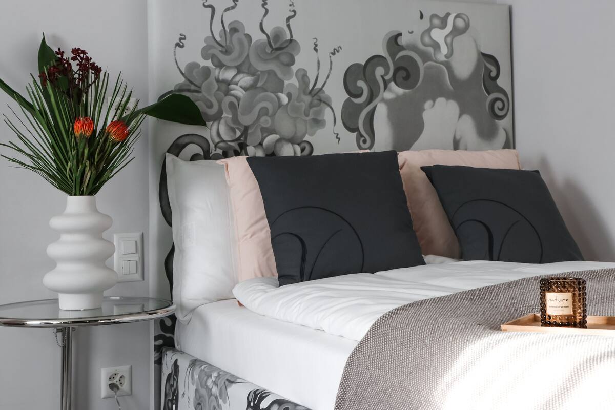 Un lit bien fait, recouvert d’une couvre-lit blanche et grise à motifs, une table d’appoint ronde sur laquelle se trouve un vase blanc contenant des feuilles vertes et des fleurs rouges, et une œuvre d’art murale aux motifs abstraits.