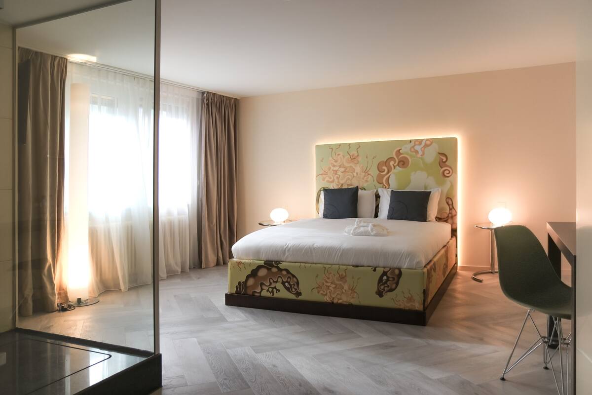 Une chambre d’hôtel avec un grand lit, un bureau et une chaise. Un miroir se trouve sur le côté gauche de la pièce.