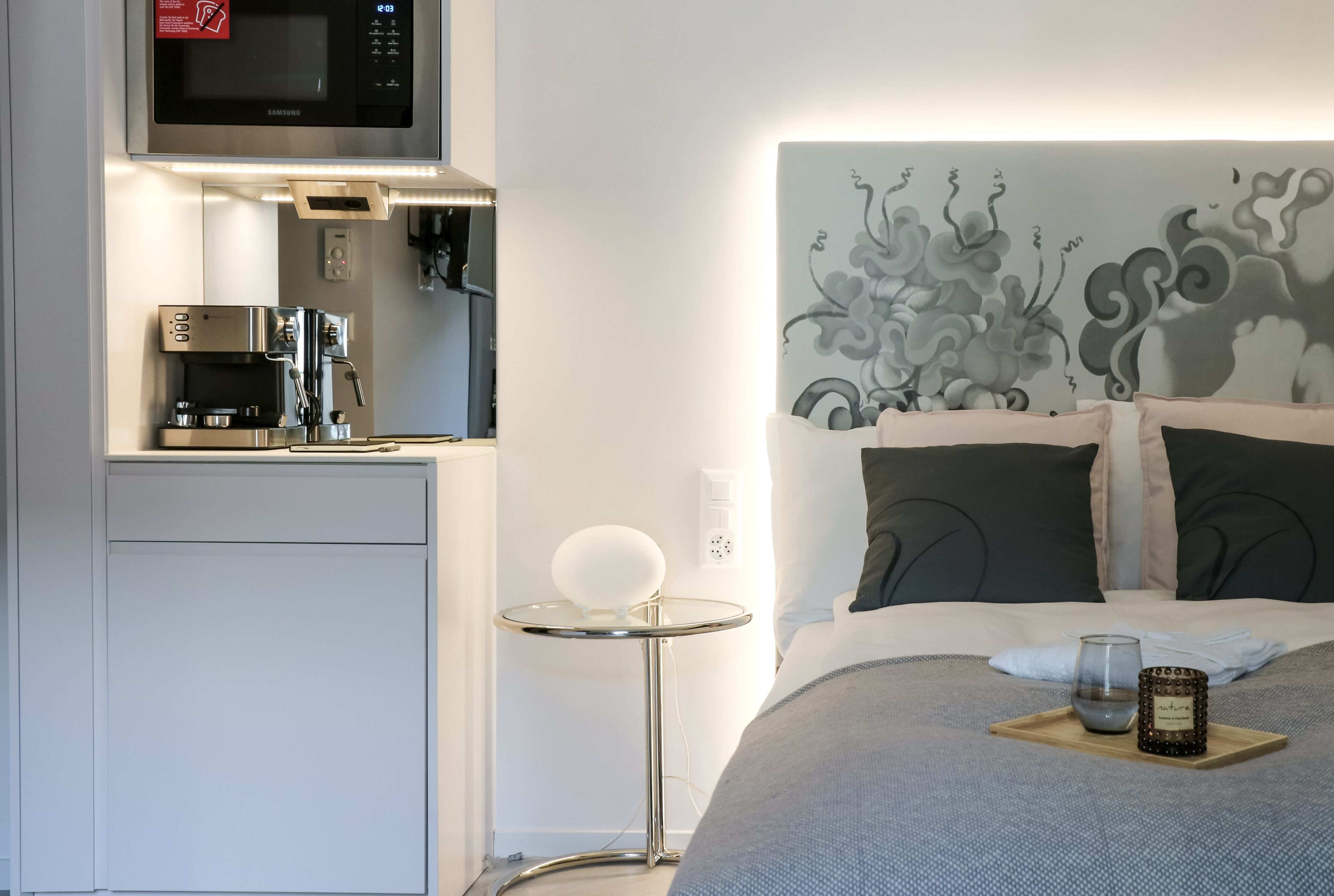 Une chambre d’hôtel compacte dotée d’une kitchenette, comprenant un four à micro-ondes, une machine à café et une petite table ronde avec une lampe blanche. Le lit est agrémenté d’une tête de lit décorative et d’un plateau avec un verre et une bougie.