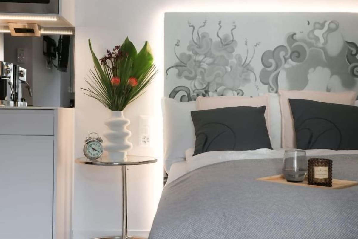 Une chambre à coucher bien rangée, avec un lit, des oreillers, une table de chevet avec une horloge et un vase rempli de fleurs.