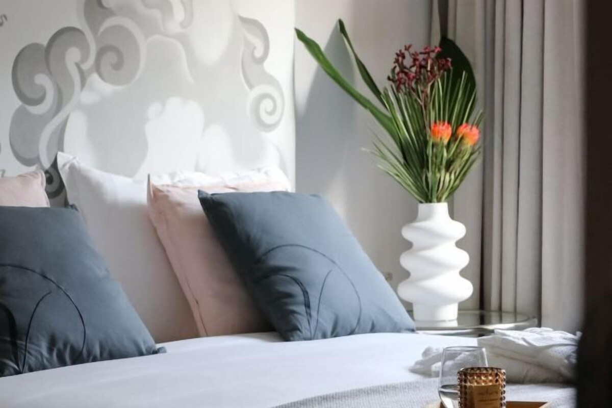 A neatly made bed with pillows, a white vase with flowers, and a bedside table with a glass and a candle.