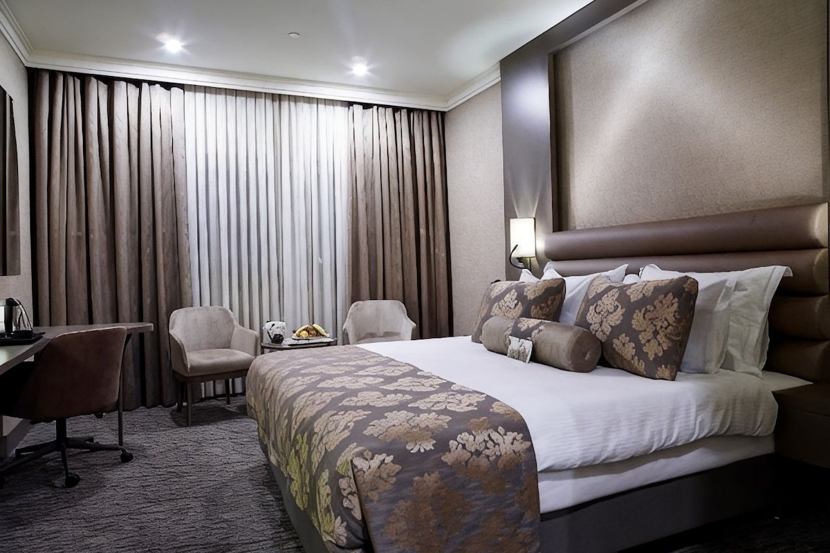 A hotel room with a bed, two chairs, a desk, and a large window with curtains.