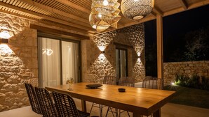 An outdoor dining area with a wooden table and wicker chairs, surrounded by stone walls and large windows. - PAPADRIA VILLAS as featured in THE TIMES - Modern Luxury Getaway nr Kathisma (Tsoukalades)