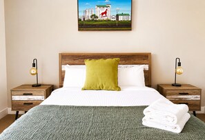 A neatly made bed with a green pillow, two bedside tables with lamps, and a painting above the bed.