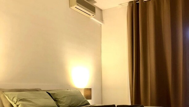 Une chambre à coucher comprenant un lit, un climatiseur fixé au mur, une lampe de chevet et une fenêtre avec des rideaux.