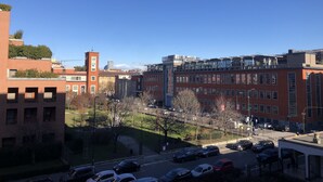 Un immeuble à plusieurs étages, dont la façade est un mélange de briques et de verre, un parking où sont garées plusieurs voitures, et un ciel bleu dégagé.