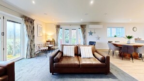 A spacious living area with a brown leather sofa, a dining table with chairs, and large windows with curtains. - Stunning SEAVIEW Coastal Cottage - Sleeping 8 in 4 Bedrooms - Pool and Beach Access Included. (Filey)