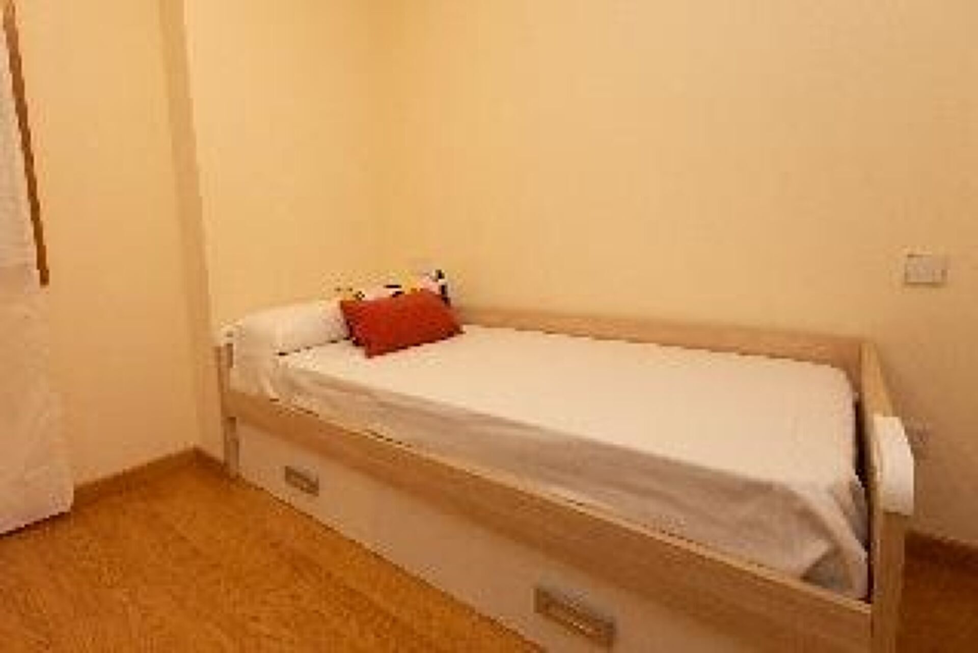 A small, single-bed room with a wooden frame, a white mattress, and a red pillow.