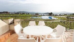 Zona de estar al aire libre con mesa y sillas, con vistas a una piscina ajardinada y montañas distantes.