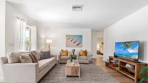Un salon avec un canapé beige, une table basse, un téléviseur et un tableau représentant une scène de plage.