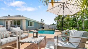 Un espace aménagé au bord de la piscine, avec des meubles d’extérieur, un parasol et une maison en arrière-plan.