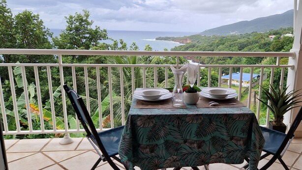 Un balcon avec une table dressée pour deux personnes, offrant une vue panoramique sur l’océan et une végétation luxuriante.