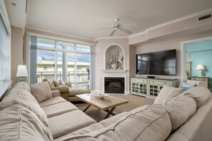 A living room with beige sofas, a wooden coffee table, a flat-screen TV, a fireplace, and a ceiling fan. - Beautiful 3 Bedroom Condo in North Ocean City with Rooftop Pool (Ocean City)