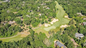 Vista aérea de un campo de golf con búnkeres de arena, rodeado de árboles densos y propiedades residenciales.