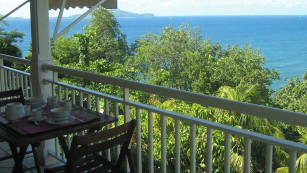 A balcony with a table set for dining, overlooking a lush landscape and the sea. - The Kolibri The Nest of Bellemont (TROIS-RIVIERES)