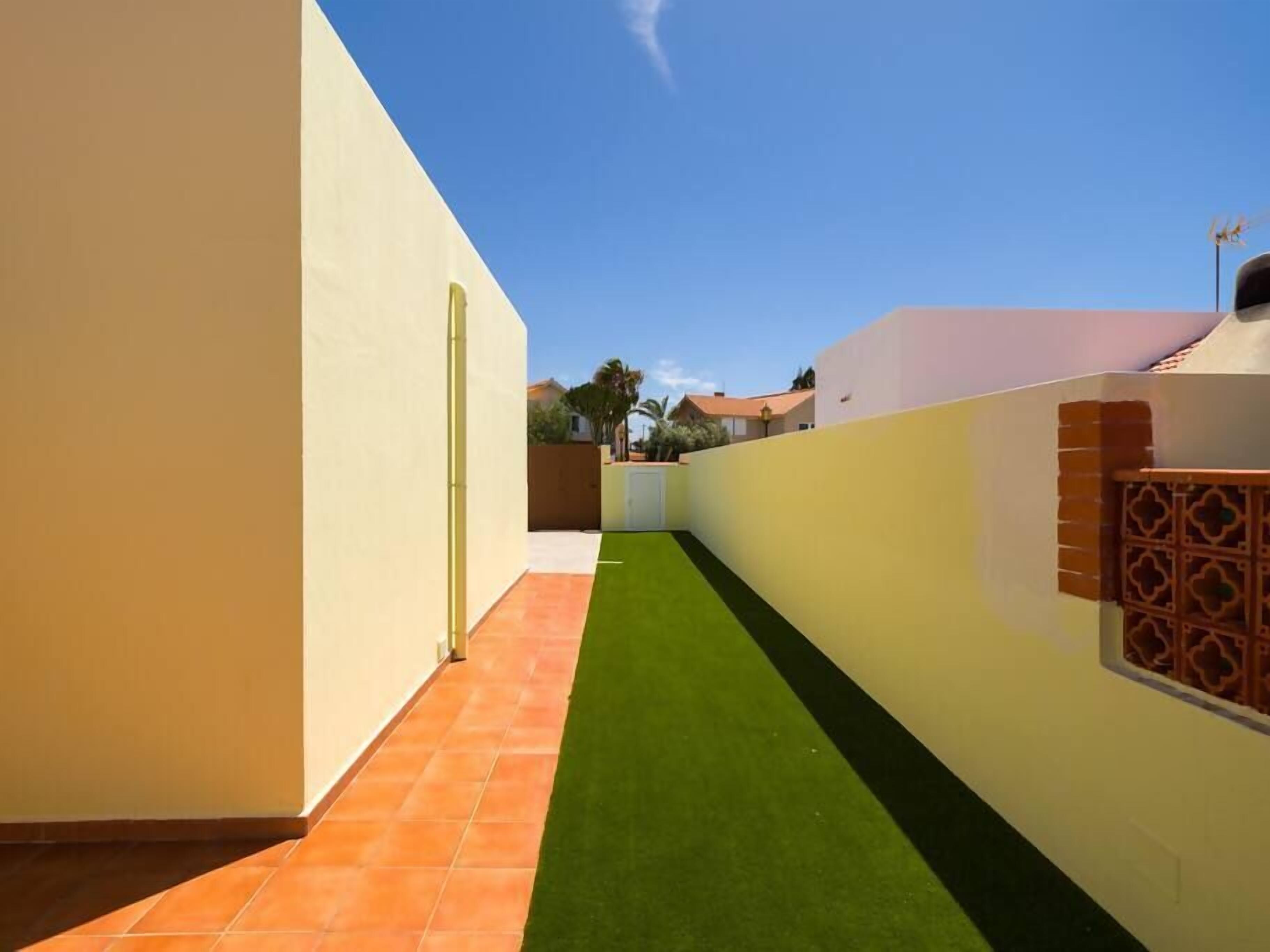 A narrow, enclosed courtyard with a green artificial turf path, a tiled floor, and a white wall with a decorative brick section.