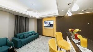 Moderni hotellihuone, jossa on turkoosi sohva, keltaiset tuolit, televisio ja hedelmäkulho juomien kera.