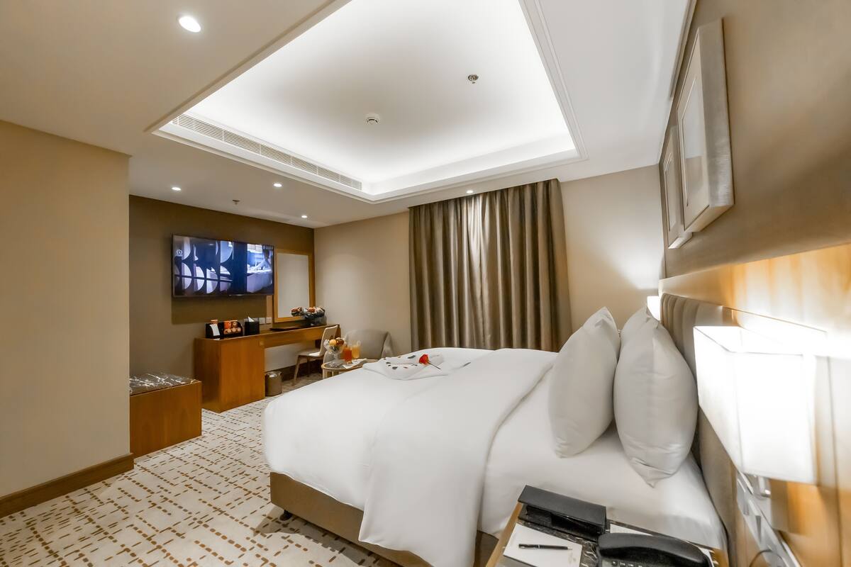 A hotel room with a large bed, a desk with a chair, a television, and a ceiling light.