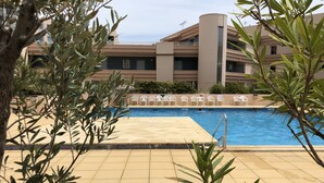 Une piscine dotée d’une terrasse carrelée, entourée de verdure et avec un immeuble à plusieurs étages en arrière-plan.