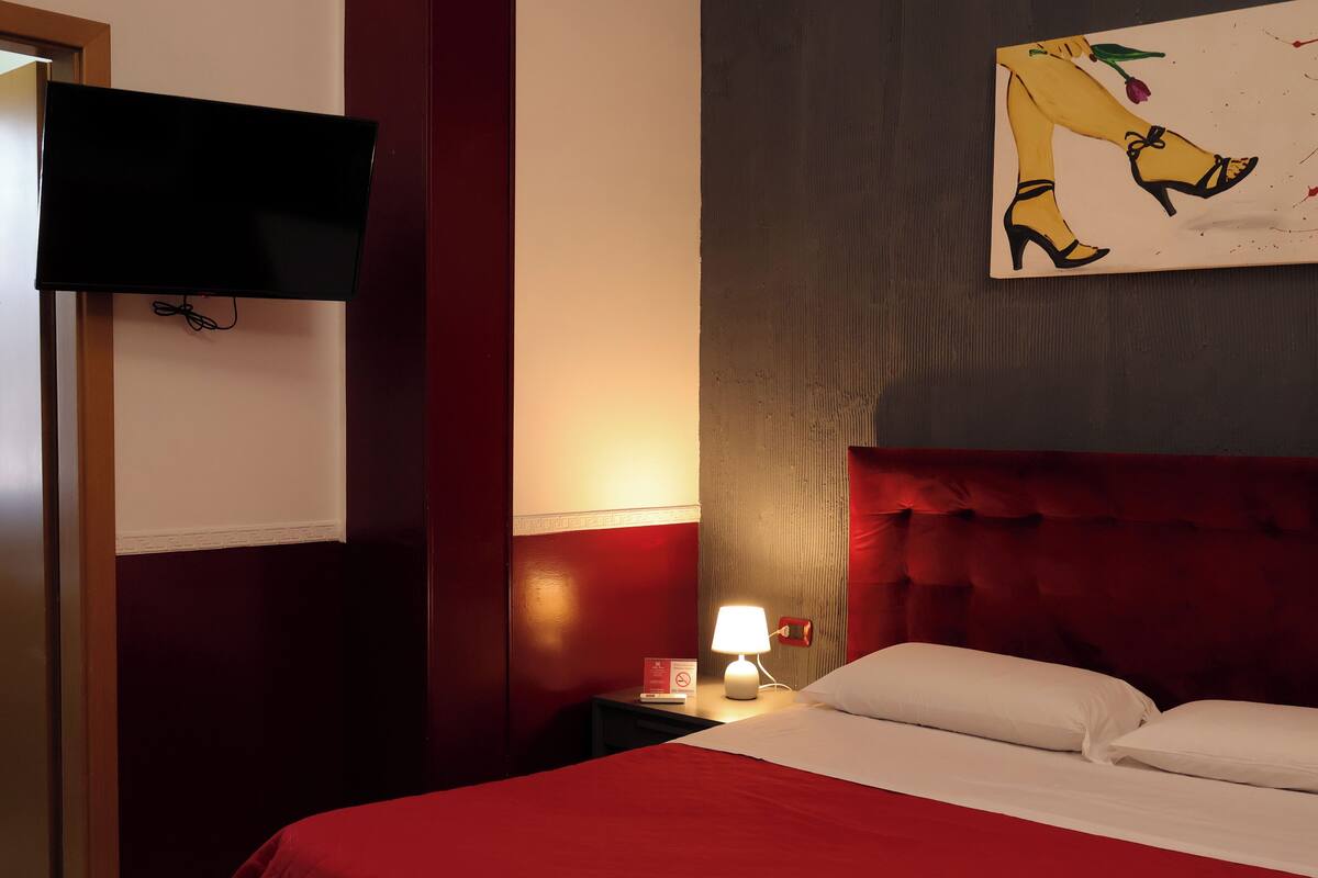 A hotel room with a bed, a TV mounted on the wall, a nightstand with a lamp, and a painting on the wall.