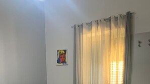 Quarto com um sofá cinza estampado, uma cama com cobertura escura e um banco. Há toalhas dobradas na cama e uma janela com cortinas leves.