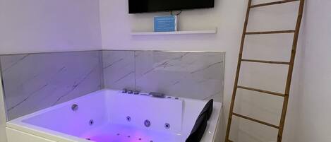 A modern bathroom with a large bathtub, a mounted television, and a wooden ladder.
