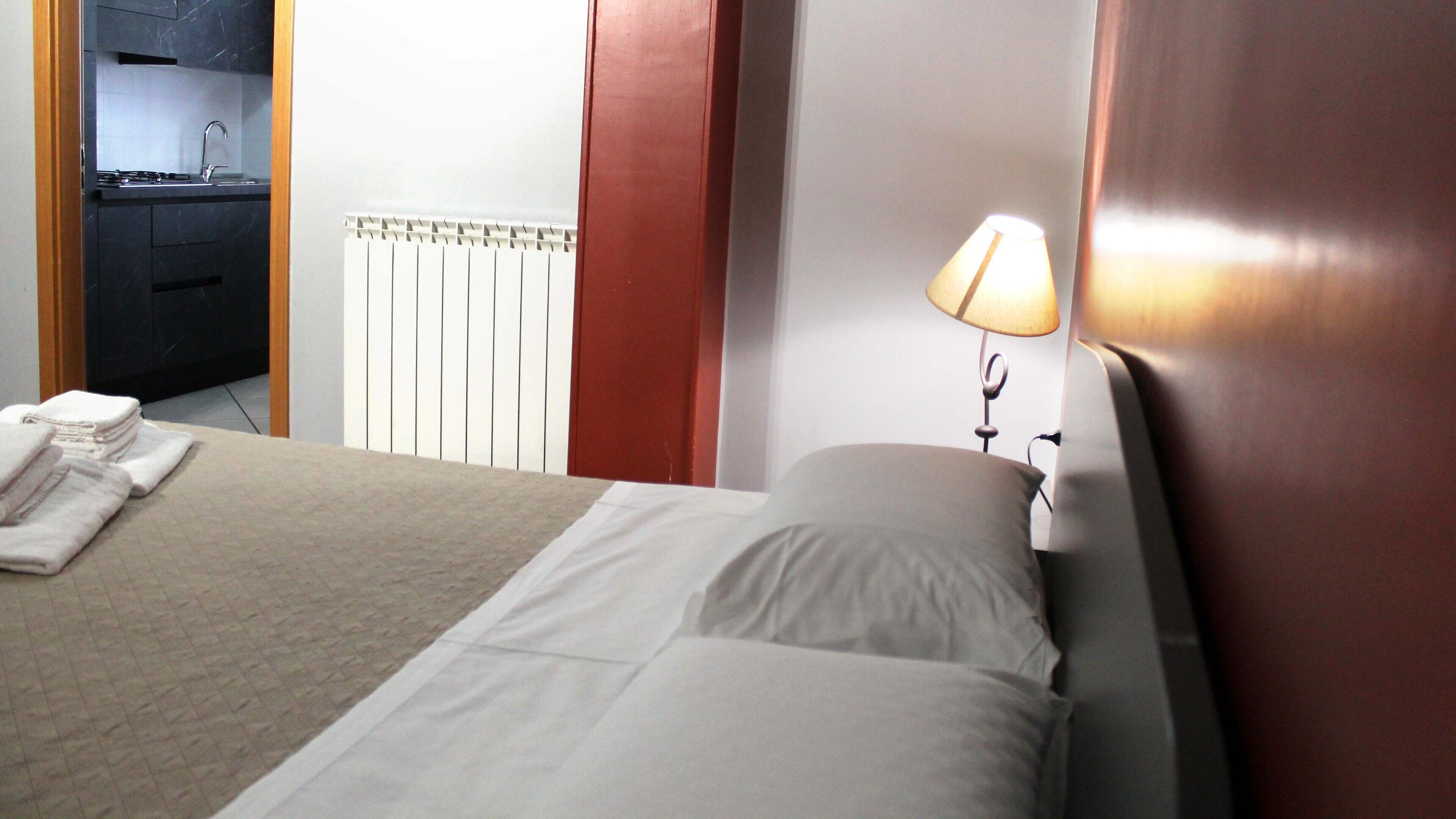 Une chambre d’hôtel comprenant un lit, une table de chevet avec une lampe, une pile de serviettes, un coin cuisine avec un évier et un radiateur.
