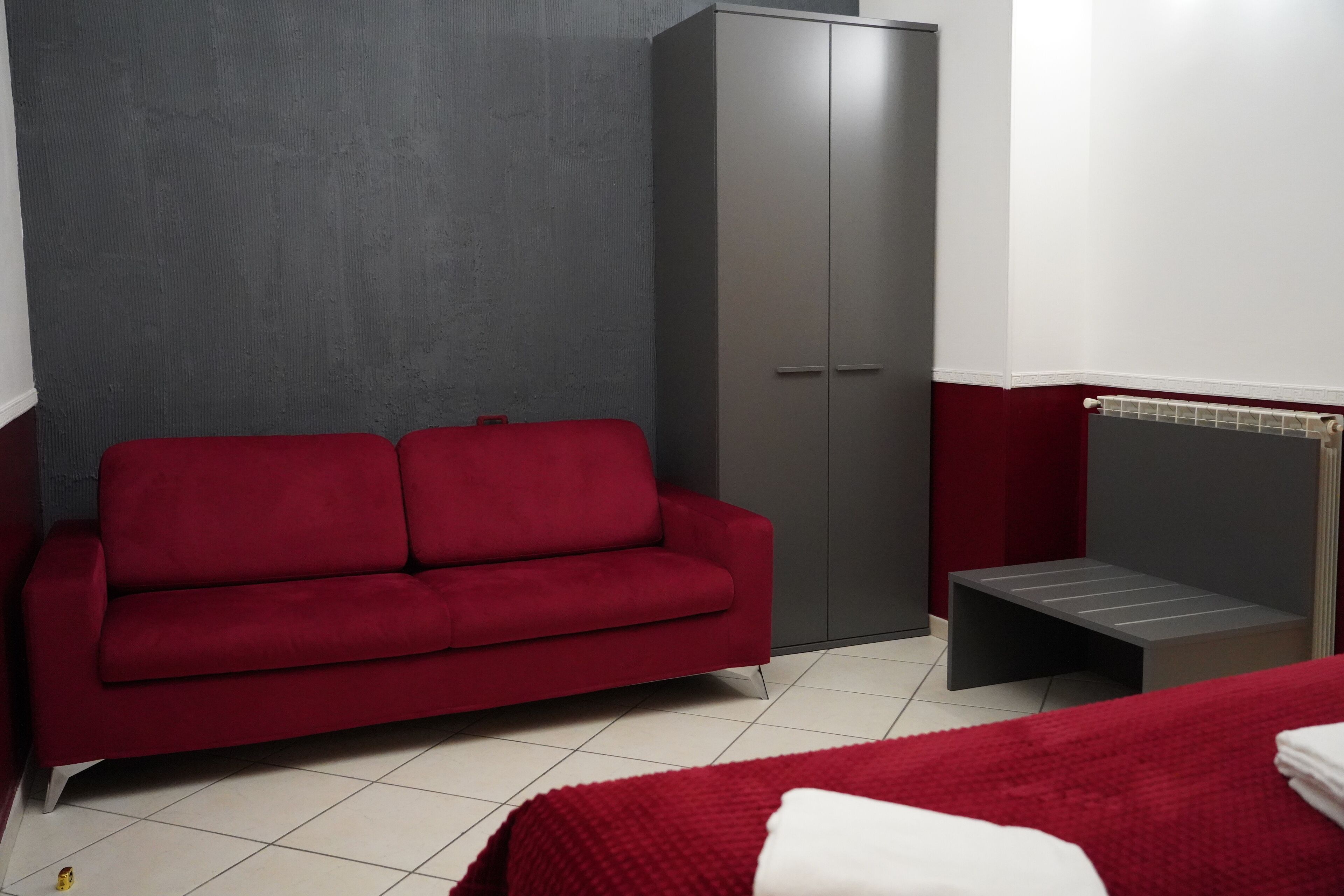 A room with a red sofa, a grey wardrobe, a small stool, and a bed with a red blanket.