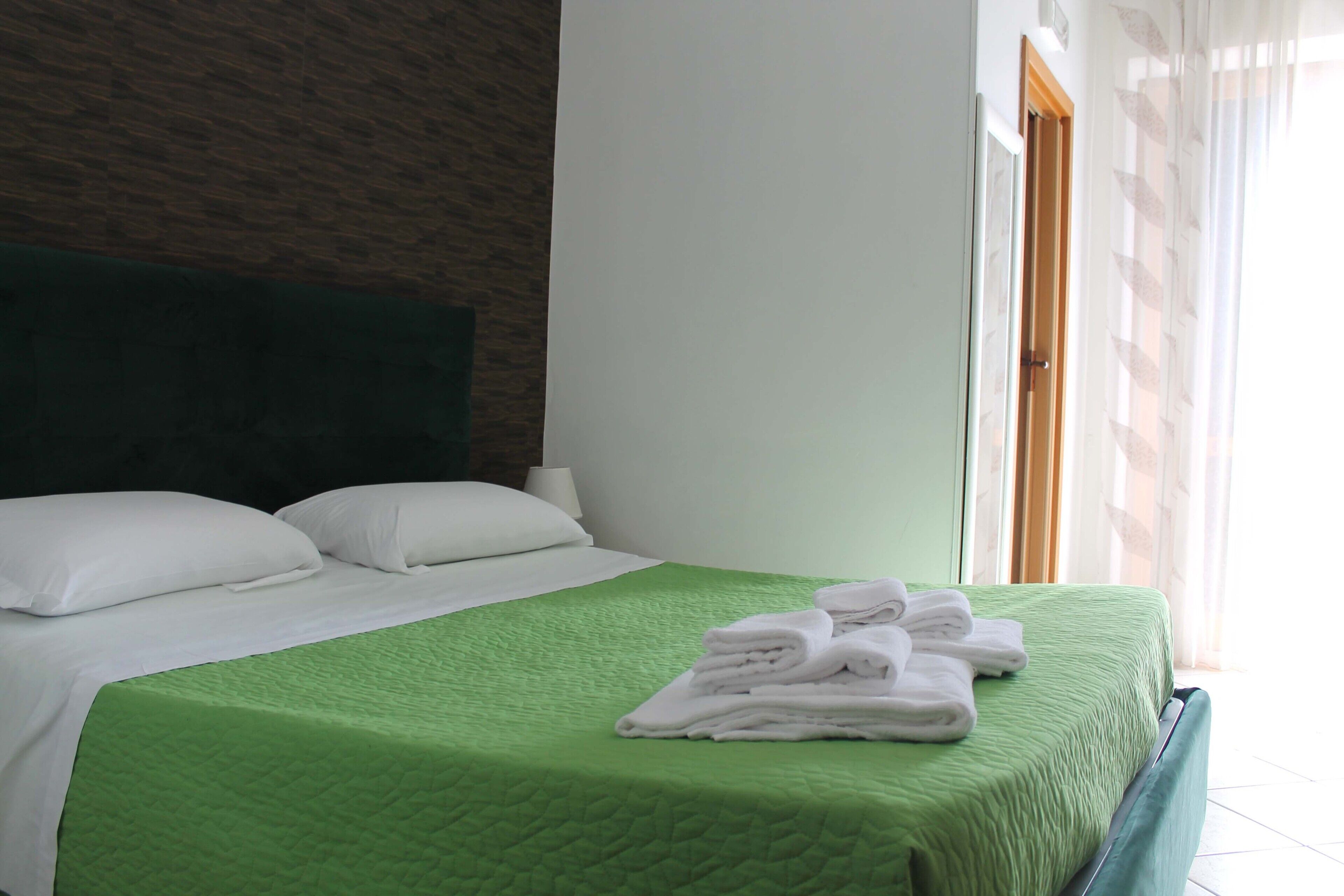 A neatly made bed with a green quilt, white pillows, and a stack of folded towels on the side.